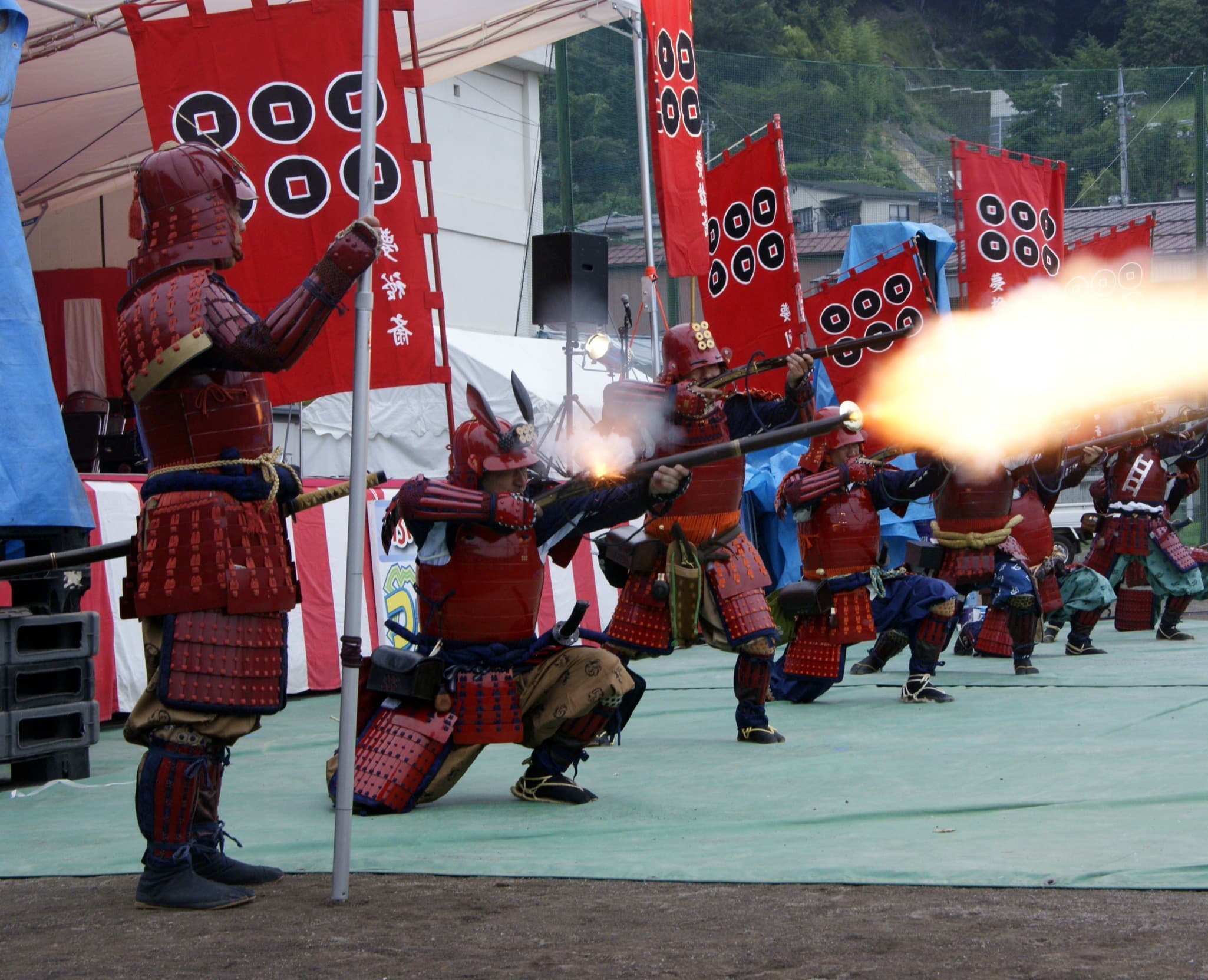 A matchlock-gun corps in red samurai armour firing a volley beneath six-coin-crest banners at a reenactment in Gunma